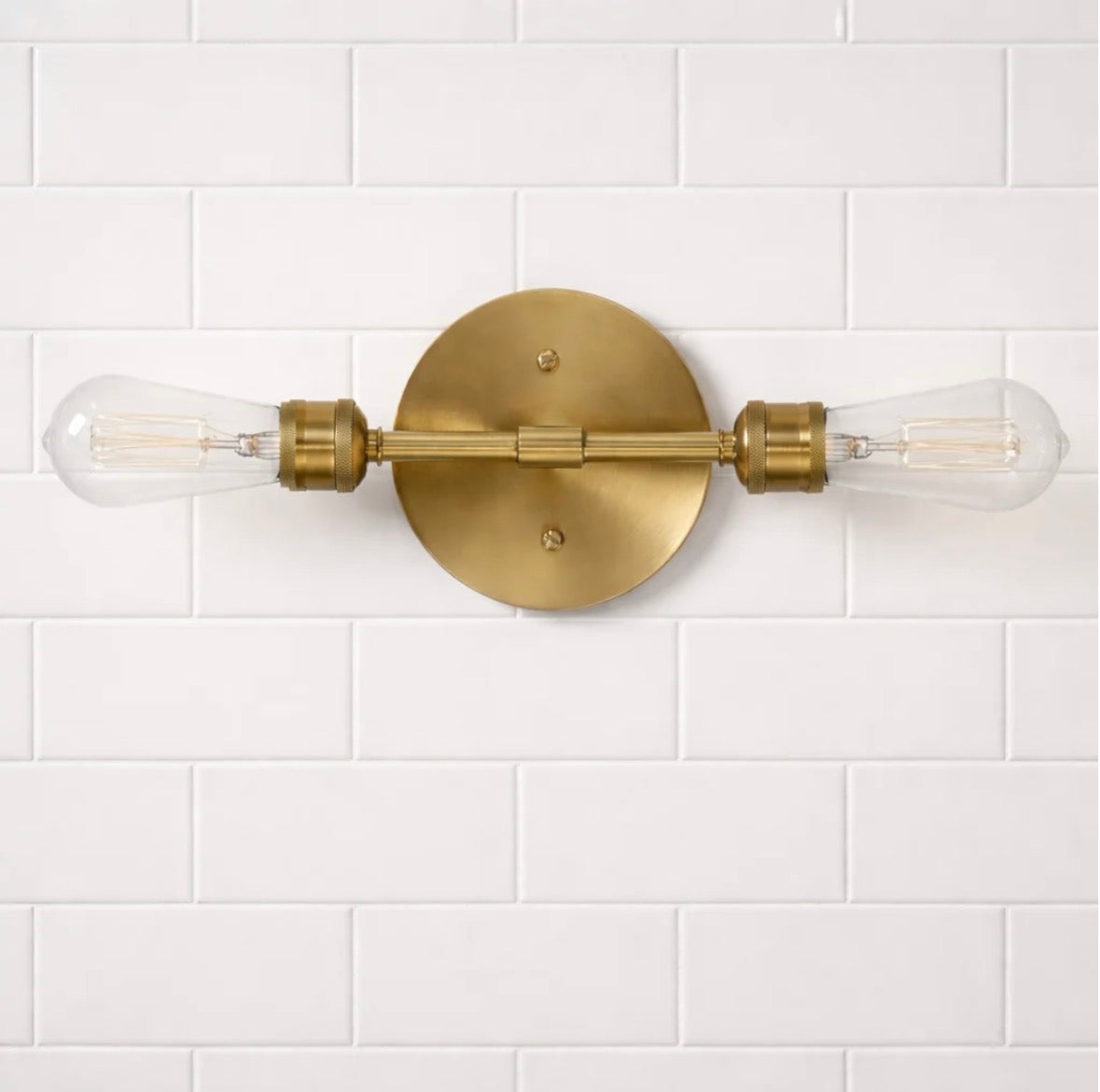 The Aurelia satin brass double-light sconce above a round mirror – warm glow lifestyle shot